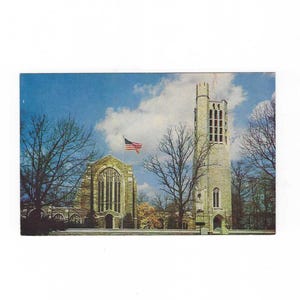 May include: Vintage postcard featuring a stone church and bell tower under a blue sky with fluffy clouds. An American flag waves atop the church. Bare trees frame the buildings, suggesting a fall or winter scene.
