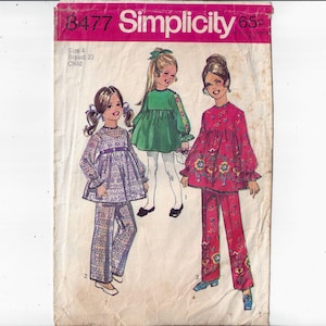 May include: Simplicity sewing pattern number 8477 for a child's dress and pants. The pattern includes three different styles: a green dress with long sleeves, a purple dress with long sleeves and pants, and a red dress with long sleeves and pants. The pattern is for size 4, with a 23-inch bust.