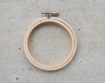 May include: A wooden embroidery hoop with a brass screw tightening mechanism. The hoop is round and has a natural wood finish.