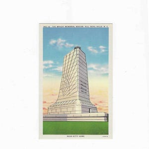 May include: Vintage postcard featuring the Wright Brothers National Memorial in Kill Devil Hills, North Carolina. The monument is a tall, white structure with a geometric design, set against a blue sky with clouds and green grass.