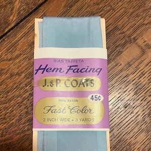Può includere: Un pacchetto di orlo in taffetà blu chiaro di J.&P. Coats. La confezione è etichettata con "Bias Taffeta Hem Facing", "J.&P. Coats", "100% Rayon", "45¢", "Fast Color" e "2 inch wide. 3 yards".