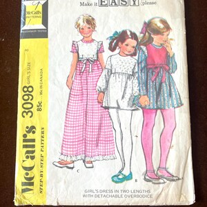 Puede incluir: Un sobre de patrón de costura McCall's 3098 para un vestido de niña en dos largos con corpiño desmontable. La portada presenta ilustraciones de tres niñas con diferentes variaciones de vestidos, incluido un vestido largo de cuadros rosas y dos estilos más cortos con telas estampadas, todos con mangas largas o cortas.