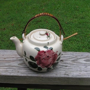 May include: A white ceramic teapot with a rose design and a bamboo handle. The teapot has a lid with a small knob and a spout. The teapot is sitting on a wooden surface.