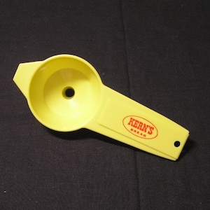 Puede incluir: Embudo de plástico amarillo con asa. El embudo tiene una abertura grande y un pico pequeño. El asa tiene la frase "Kern's Bread" impresa.