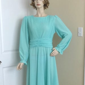 May include: A light blue pleated dress with long sleeves and a gathered waistline. The dress is on a mannequin in a room with white trim.
