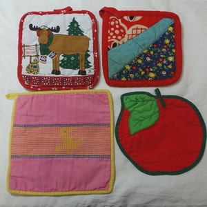 May include: Four colourful pot holders. One features a moose with a scarf and the words "Moose Crossing." Another has a floral pattern, and a third is pink with a yellow embroidered design. The last is an apple shape.