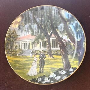 May include: Decorative plate featuring a painted scene of a couple in period clothing standing in front of a large house with a red roof. The plate has a gold rim and a lush green lawn with white flowers.
