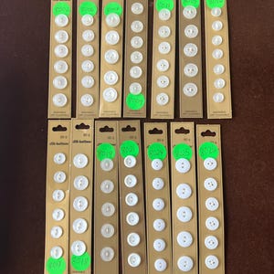 May include: A collection of white, round buttons in various sizes, displayed on brown cardboard packaging. Each card has multiple buttons, with the text "dill-buttons" and "WASHABLE DRY CLEANABLE".