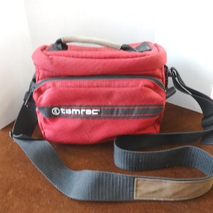May include: A red Tamrac camera bag with a black strap and handle. The bag has a front pocket and the Tamrac logo in white. The shoulder strap is black and has a brown pad for comfort.