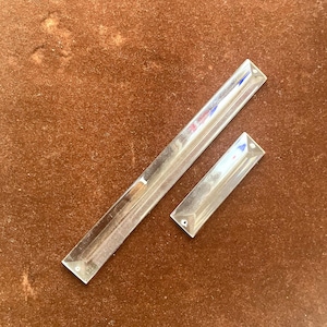 Puede incluir: Dos prismas de cristal rectangulares y transparentes con bordes biselados. Un prisma es más largo que el otro. Cada uno tiene un pequeño agujero en cada extremo. Los prismas se muestran sobre una superficie marrón.