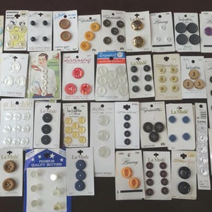 Peut inclure: Une collection de boutons variés sur cartes, de différentes couleurs, tailles et matériaux. Certaines cartes comportent du texte comme "La Mode" et "Premium Quality Buttons". Les boutons sont disposés sur une surface marron.