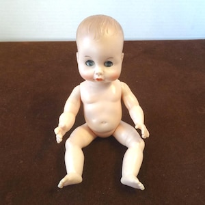 Può includere: Una bambola vintage con un viso dipinto, occhi blu e un leggero sorriso. La bambola ha un incarnato chiaro, capelli modellati ed è posizionata seduta con le braccia e le gambe distese. La bambola è realizzata in plastica dura.