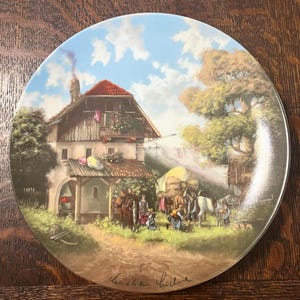 以下が含まれることがあります： 田園風景の詳細な絵画が施された装飾皿。アートワークは、赤い屋根の2階建ての建物と、木々や人物に囲まれた様子を描いています。このお皿は光沢のある仕上がりで、茶色の縁取りが施されています。