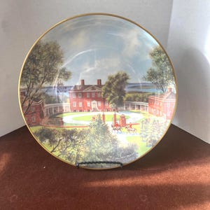 May include: Decorative plate with a gold rim featuring a detailed landscape scene. The image depicts a red brick building with a white fence, trees, and figures on horseback. The plate has a glossy finish.