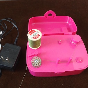 May include: A bright pink portable sewing machine with a spool of green thread and a bobbin. The machine is open, revealing its interior components. A black power adapter and cord are connected to the machine.