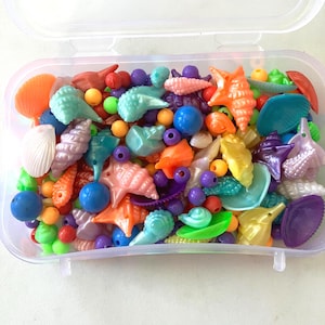 May include: A clear plastic rectangular storage container filled with a variety of colorful plastic beads shaped like seashells, starfish, and small spheres in shades of orange, blue, green, purple, yellow, and white.