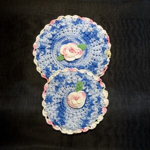 May include: Two crocheted doilies with a blue and white color scheme. Each doily features a pink flower in the center.
