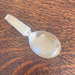 May include: A silver spoon with a rounded bowl and a rectangular handle. The handle has a decorative design near the bowl. The spoon is set against a dark wood surface.