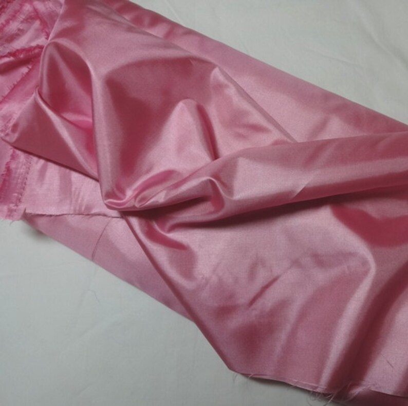 May include: A swatch of pink satin fabric, with a lustrous sheen, is draped across a surface. The fabric is a solid, light pink color, and the edges are unfinished. The folds and creases in the fabric create shadows and highlights.