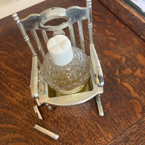 May include: A small, clear glass perfume bottle with a white cap sits in a miniature silver rocking chair. The bottle has a textured, bubble-like pattern. The chair has a broken piece on the left side. The background is a wooden surface.