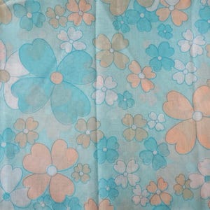 May include: A fabric with a repeating floral pattern. The flowers are in shades of blue, peach, white, and tan, set against a light blue background. The flowers have a retro, stylized design, with rounded petals and a central dot.