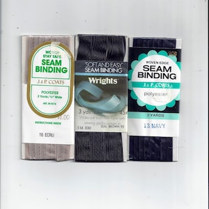 May include: Three rolls of seam binding tape. The first roll is white and says "Woven Stay Tape Seam Binding J.& P. Coats Polyester 3 Yards 1/2" Wide Art. M.9274 1.00 16 Ecru". The second roll is black and says "Soft and Easy Seam Binding Wrights 3 yds. 2.75m 45¢ 116 330 Wider soft finish hem tape with sewing guides woven in Seal Brown 92". The third roll is navy blue and says "Woven Edge 80¢ Seam Binding J.& P. Coats polyester 3 Yards 13 Navy".
