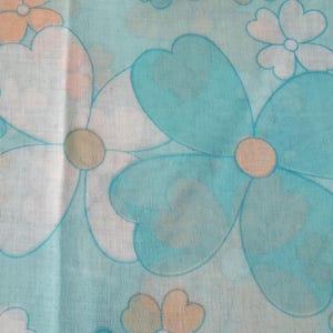 May include: Close-up of a fabric with a repeating floral pattern. Large flowers in shades of blue, white, and peach are scattered across a light blue background. The flowers have a retro, 1970s style, with rounded petals and a simple design.