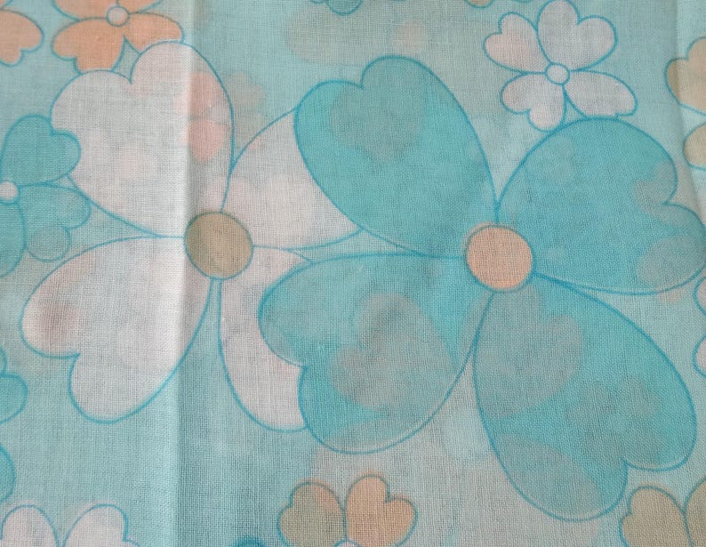 May include: Close-up of a fabric with a repeating floral pattern. Large flowers in shades of blue, white, and peach are scattered across a light blue background. The flowers have a retro, 1970s style, with rounded petals and a simple design.