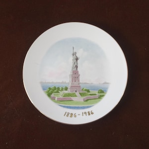 May include: A white ceramic plate with a gold rim features a colorful illustration of the Statue of Liberty on a green island. The plate has the text "1886-1986" in gold lettering.