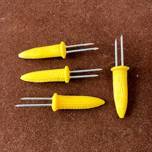May include: Four yellow corn-shaped corn holders with stainless steel prongs. Three holders have two prongs, and one has two prongs. The holders are made of yellow plastic and are designed to resemble miniature corn cobs.