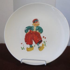 May include: White ceramic plate with a colorful illustration of a boy wearing red pants and wooden shoes. The boy is standing in a field of green grass and red flowers.