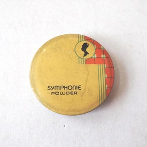 May include: A vintage, round Symphonie Powder compact with a yellow lid. The lid features the text "SYMPHONIE POWDER" and a silhouette of a woman's profile in a circle, with a red and white plaid design on the right side.