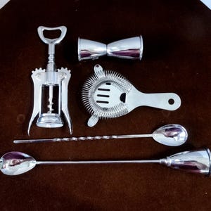 Puede incluir: Un conjunto de herramientas de bar plateadas que incluyen un sacacorchos, un medidor doble, un colador y dos cucharas de bar. Las herramientas están dispuestas sobre una superficie marrón, para cócteles.