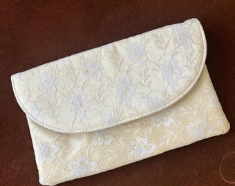 May include: Ivory-colored clutch with a floral lace pattern. The flap-over style bag is made of a soft fabric and has a rounded top. The clutch is a light cream color with a delicate, repeating floral design.
