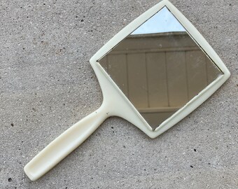 May include: A white plastic hand mirror with a square-shaped reflective surface and a long handle.