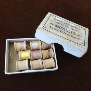 May include: A vintage box of sewing silk thread spools. The box is open, revealing a dozen wooden spools in assorted colors. The box reads "ONE DOZEN SPOOLS SEWING SILK MIDGETS ASSORTED COLORS."