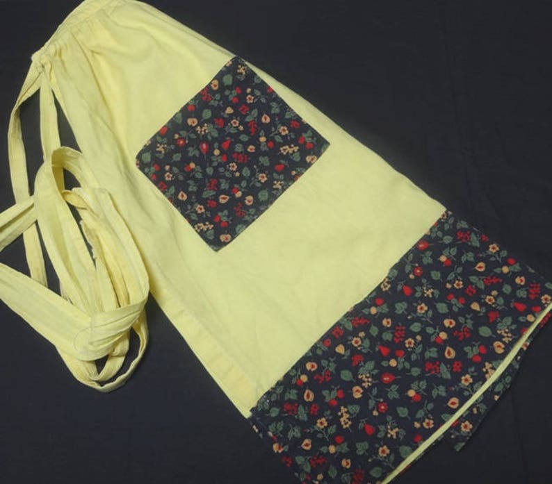May include: A yellow apron with a black and white floral print. The apron has a large pocket on the front and ties at the waist.