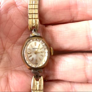 May include: A vintage gold-toned wristwatch with a small oval face. The watch face is a light colour with gold-toned hands and markers. The watch strap is also gold-toned and has a delicate, linked design.