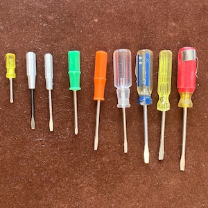 May include: A collection of eight screwdrivers with various colored handles, including yellow, white, green, orange, clear, blue, and red. The screwdrivers are arranged in a row, showcasing their different sizes and designs.