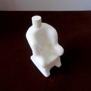May include: White ceramic bottle shaped like a rocking chair. The bottle has a white plastic lid.
