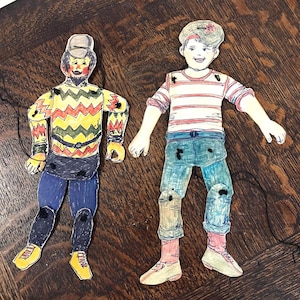 May include: Two handmade paper puppets. One puppet has a yellow, red, and green patterned shirt, blue pants, and a gray hat. The other puppet has a red and white striped shirt, blue jeans, and a gray hat with a flower.