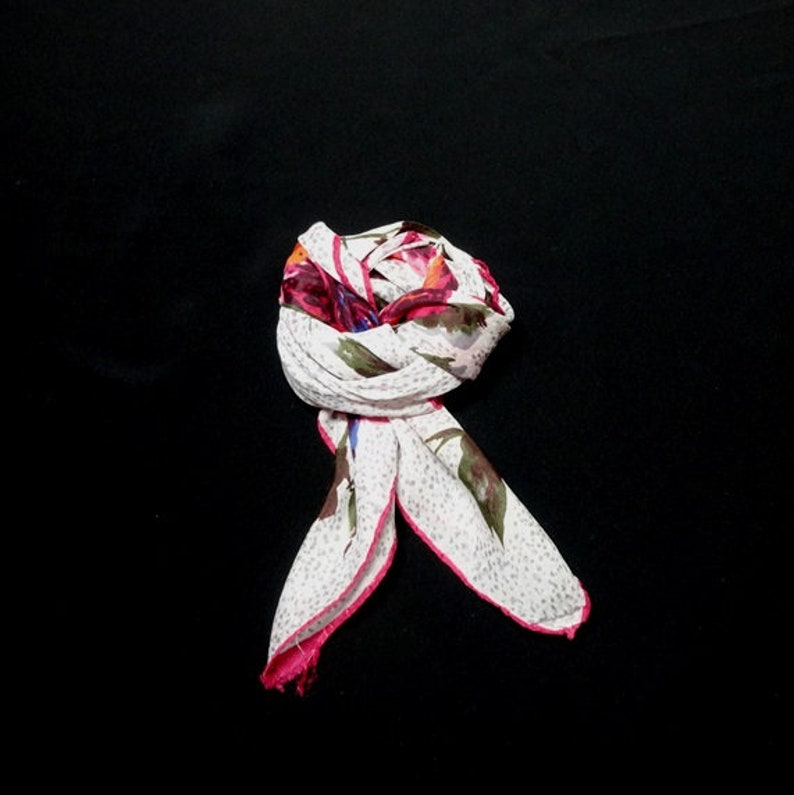 May include: A white scarf with a pink border and a floral print in shades of pink, blue, and green. The scarf is folded and tied into a knot.
