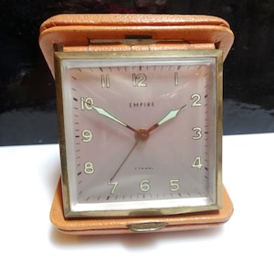 May include: A vintage Empire travel clock in a tan leather case. The clock face is a light pink with black numerals and gold-tone hands. The clock is rectangular with a gold-tone frame and a hinged leather case.