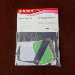 May include: A Singer Iron-On Repair Kit in a clear plastic package. The kit includes various fabric patches in green, gray, and white, with a dark blue accent. The packaging features the Singer logo and text in multiple languages.