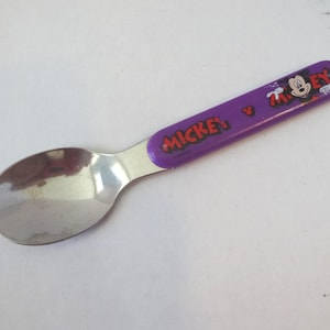May include: A stainless steel spoon with a purple handle featuring the word "Mickey" and a cartoon image of Mickey Mouse. The spoon has a rounded bowl and is designed for children.