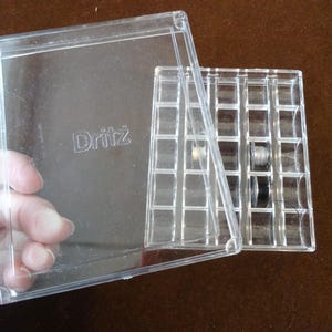 Puede incluir: Un organizador de hilo Dritz de plástico transparente con una tapa abierta. El organizador tiene una cuadrícula de pequeños compartimentos, algunos con carretes de hilo de varios colores. La palabra "Dritz" está impresa en la tapa.