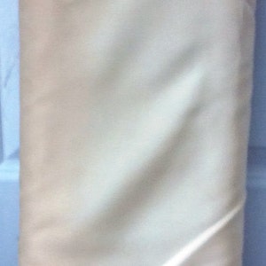 May include: A tightly rolled bundle of fabric, primarily cream-colored with subtle shading and folds. The bottom edge has a warm, tan hue. The fabric appears to be a soft, possibly natural material, suitable for sewing or crafting projects.