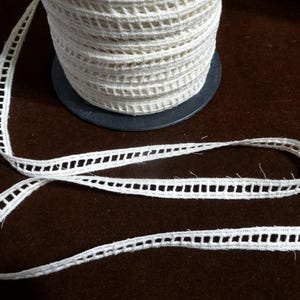 May include: A spool of white cotton crochet trim with a delicate openwork design. The trim is approximately 1/2 inch wide and has a scalloped edge.
