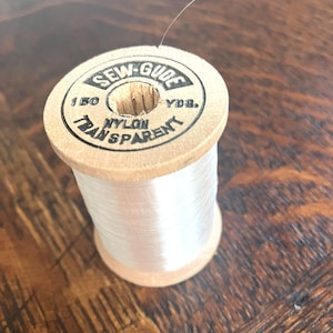 Puede incluir: Un carrete de hilo de nailon transparente Sew-Gude con un núcleo de madera clara. La etiqueta superior muestra el texto Sew-Gude, 150 Yds. y Nylon Transparent en negro. El hilo es transparente y está enrollado en el cuerpo cilíndrico.