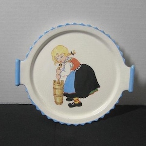 Könnte beinhalten: Ein weißer Keramikteller mit blauem Rand und einem Mädchen in einem blau-schwarzen Kleid, das Butter rührt. Der Teller hat zwei blaue Griffe.